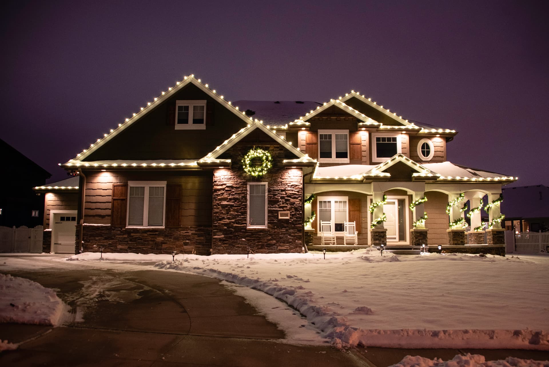 Evening residential exterior with warm white roofline lighting, an illuminated wreath, greenery, and lit porch columns